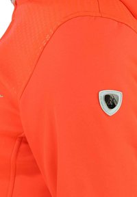 Veste orange vif avec un panneau d'épaule texturé. Présente un patch logo hexagonal en blanc et noir sur la manche. Finition en tissu lisse.