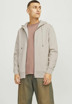 Light gray zip-up hoodie with two front pockets, drawstring hood, and ribbed cuffs, layered over a mauve shirt and paired with relaxed-fit pants.