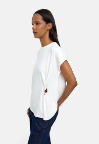 Biała bluzka z krótkim rękawem o luźnym kroju, z ozdobnym charmsiem z czerwonych i białych koralików przymocowanym do szwu bocznego.