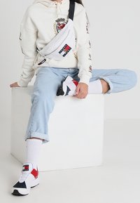 Borsa a tracolla bianca con logo, jeans azzurri arrotolati, calzini bianchi, sneakers rosse e blu, e una felpa beige con dettagli dell'emblema.