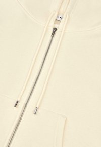 Sudadera cremosa con cremallera, hecha de un tejido suave, que cuenta con un bolsillo frontal, cordones y detalles de cremallera plateada. Diseño minimalista con líneas limpias.