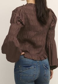 Blusa marrone con arricciature, maniche lunghe a sbuffo e orlo arricciato. Presenta una superficie strutturata e un colletto alto. Abbinata a jeans attillati blu.