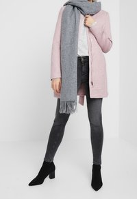 Lichtroze jas met knopen, grijze sjaal met franjes, grijze jeans en zwarte enkellaarsjes. Gelaagde materialen en een gestructureerd ontwerp.