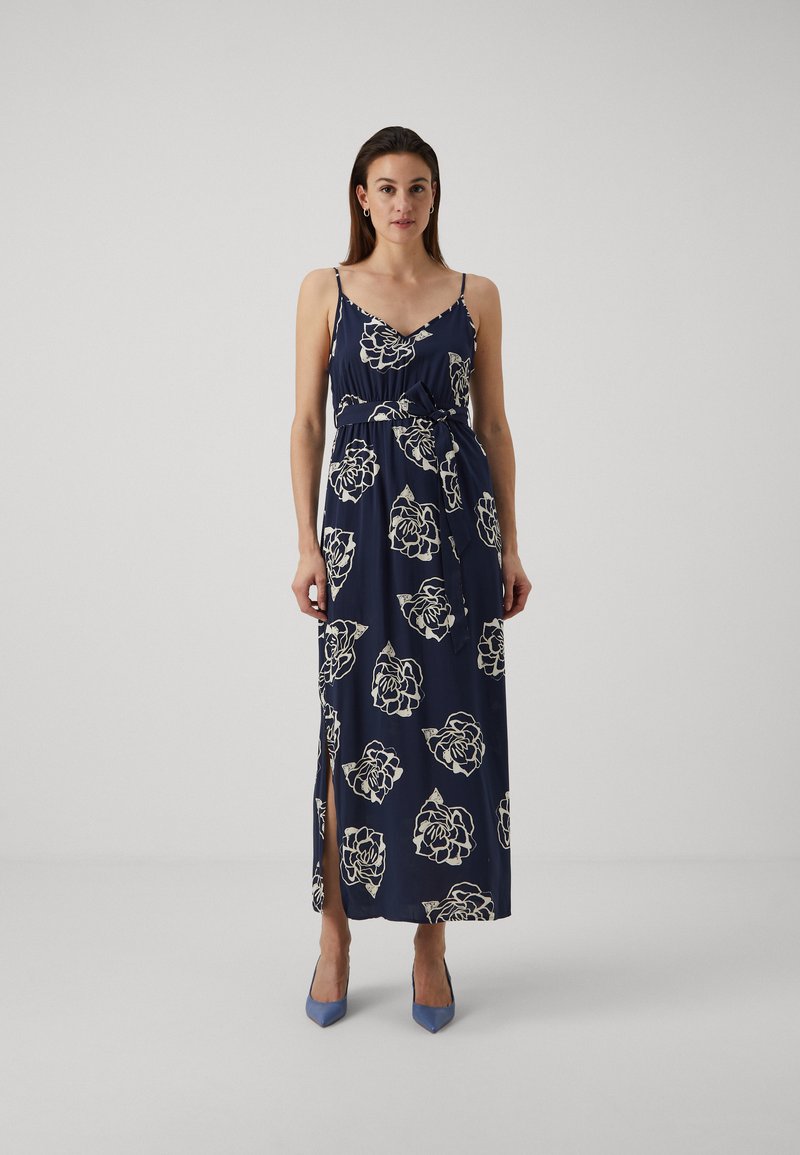 Robe maxi marine avec des motifs floraux blancs, fines bretelles, corsage froncé, ceinture à la taille et fente sur le côté, associée à des talons bleu clair.