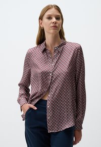 Langärmliges Hemd mit einem rot-blauen geometrischen Muster, aus glattem Stoff gefertigt, mit Kragen und Knopfleiste, in dunkle Hose gesteckt.