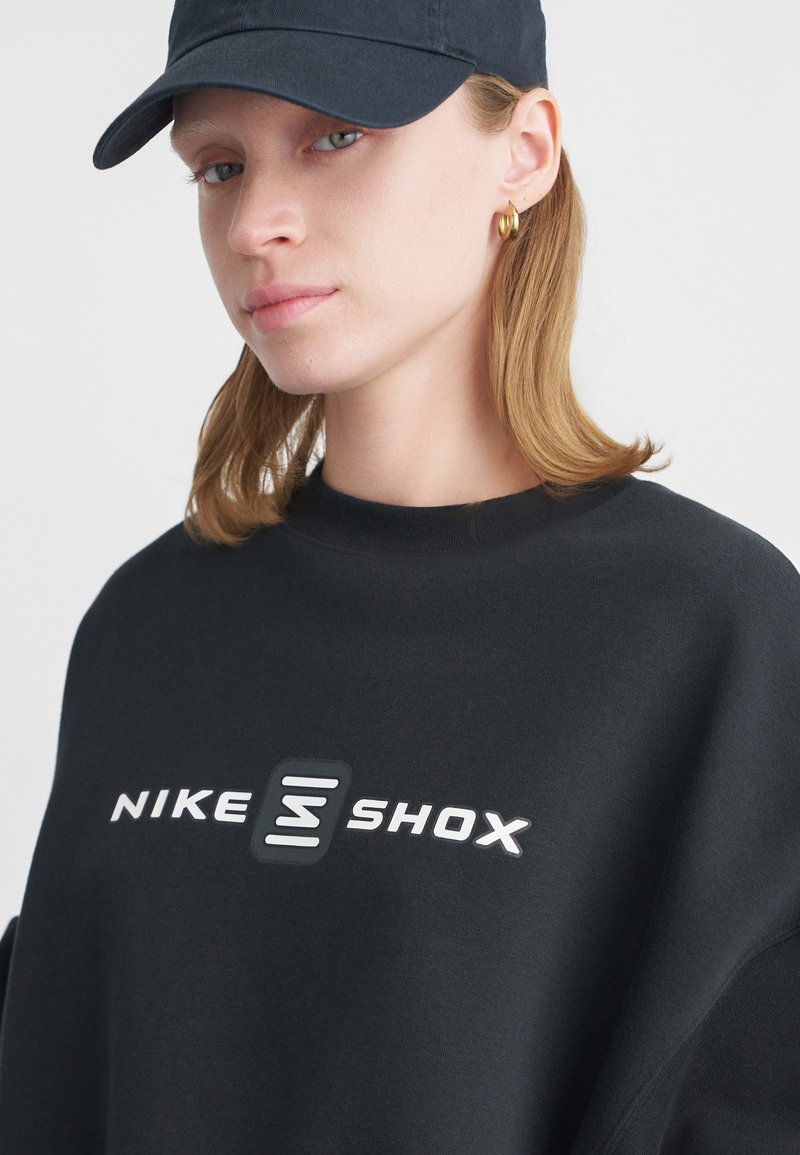 Černá mikina s vysokým límcem a velkým bílým logem "NIKE SHOX" na přední straně, doplněná tmavou čepicí a zlatými kruhovými náušnicemi.