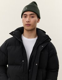 Giovane vestito con un piumino nero sopra una camicia bianca e un berretto di maglia verde oliva, in piedi contro uno sfondo chiaro e uniforme.
