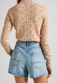 Sweter w kolorze brzoskwiniowym, dziergany, z wzorami i ściągaczem przy kołnierzu, połączony z jasnoniebieskimi szortami dżinsowymi z postrzępionymi brzegami i dwiema tylnymi kieszeniami.