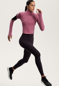 Sportjakke i mørkerød, høj krave, lynlås lukning, lange ærmer. Sorte leggings, tætsiddende, med geometriske mønstre. Sorte løbesko.