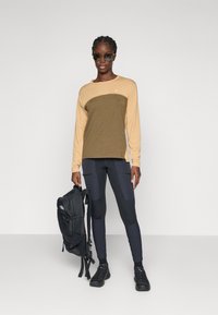 Vrouw met zonnebril, beige en olijfgroene lange mouwen shirt, zwarte leggings en sneakers, die een zwarte rugzak naast zich vasthoudt.