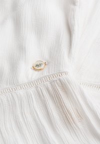 Witte, gestructureerde stof met geplooide stiksels en een klein rond goud-wit label met het opschrift "Superdry" eraan bevestigd.