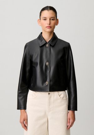 Femme aux cheveux foncés attachés en un chignon lisse, portant une veste en cuir noire boutonnée et un pantalon blanc taille haute, sur un fond gris uni.