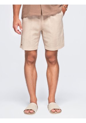 Hombre con pantalones cortos beige, sandalias tipo slide a juego y una camisa marrón, de pie frente a un fondo liso.