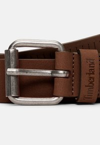 Ceinture en cuir marron avec une large boucle en métal argenté. Présente des coutures et une étiquette de marque, mettant en valeur une surface texturée et des trous ajustables.
