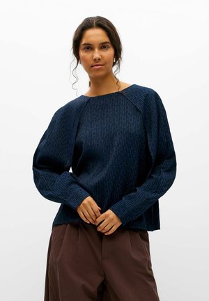 Une femme porte un chemisier bleu marine à motifs, aux manches volumineuses, et un pantalon marron sur un fond blanc uni.