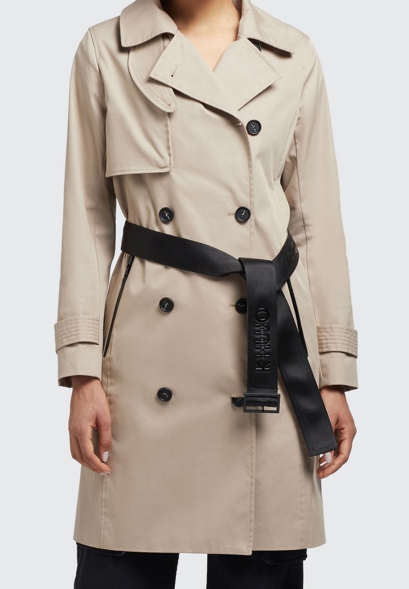 Beige trenchcoat med dubbelknäppning, svarta knappar och ett brett svart bälte med varumärke. Slät textur och märkt krage.