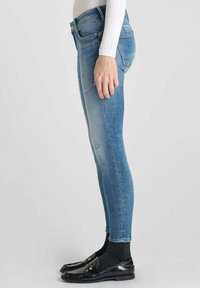 Jean skinny en denim bleu avec une finition décolorée, cinq poches et un léger effet usé. Portés avec des chaussures noires brillantes et des chaussettes noires.