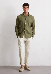 Lightweight olive green linen shirt with a button front and chest pocket, paired with cream-colored pants and beige shoes. Neutral textures.