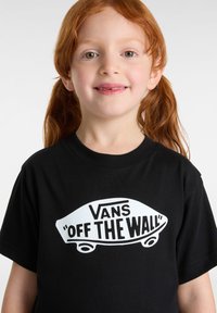 Dívka s červenými vlasy v copáncích se usmívá, má na sobě černé tričko Vans "Off The Wall" s bílým logem skateboardu na jednoduchém pozadí.