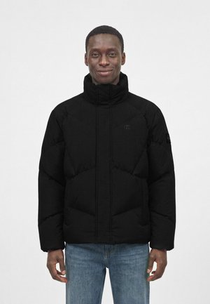 Mand iført en sort dunjakke med høj krave og blå jeans, stående mod en ensartet lys baggrund.