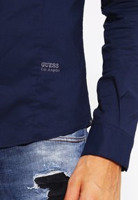Zbliżenie osoby w granatowej koszulce Guess Los Angeles oraz w podartych niebieskich dżinsach z czarną łatą tkaninową.