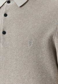 Beige gebreide poloshirt met een gestructureerde stof, drie zwarte knopen en een subtiele logo in een lichtere tint op de linkerborst.