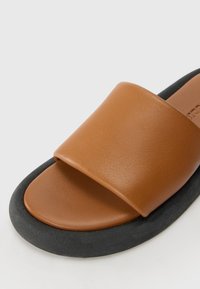 Sandale à enfiler en cuir marron avec une large bride et une semelle noire rembourrée, présentée en gros plan sur un fond uni.