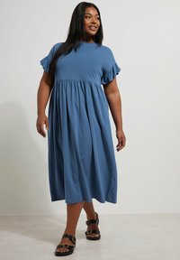 Niebieska bawełniana sukienka z krótkimi falbaniastymi rękawami, zebranym pasem i długości do połowy łydki. Połączona z czarnymi sandałami z podwójnymi klamrami.