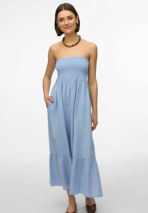 Femme portant une robe longue bleu clair sans bretelles avec un ourlet à volants, des chaussures marron et un gros collier de perles marron, debout avec une main dans la poche.