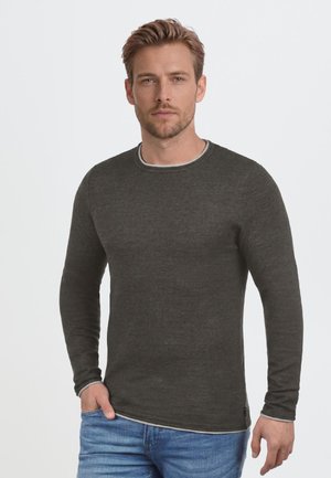 Homme aux cheveux châtain clair portant une chemise à manches longues gris foncé et un jean bleu, debout devant un fond clair uni.