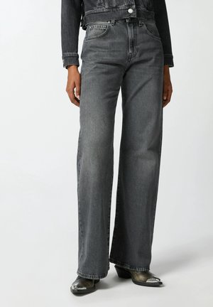 Osoba ubrana w czarne wyblakłe dżinsy z wysokim stanem i szerokimi nogawkami, czarną kurtkę dżinsową oraz czarne botki na ostro zakończonych noskach z srebrnymi czubkami.