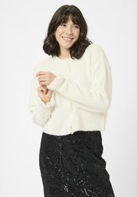 Witte, pluizige korte cardigan met een ronde halslijn en kleine knopen, gecombineerd met een zwarte paillettenrok met geometrische patronen.