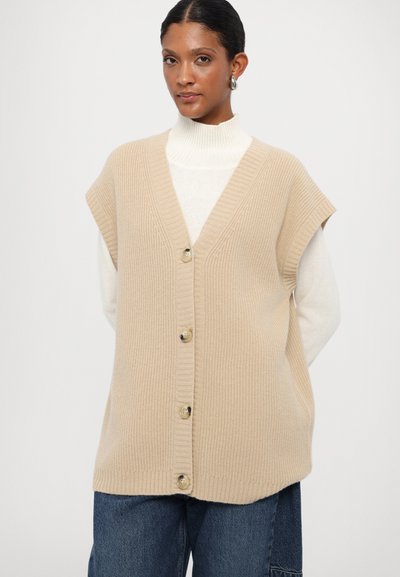 Beige strikvest med V-hals, korte ærmer og fem knapper foran. Båret over en hvid rollkrave og parret med blå jeans.