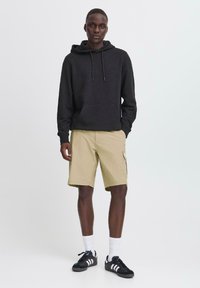 Sort hættetrøje med snore, parret med beige cargo shorts. Modellen bærer sorte træningssko og hvide sokker. Simpelt design, afslappet look.