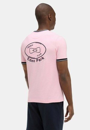 Homme portant un polo à manches courtes rose clair avec bordure noire et blanche, logo de nœud papillon noir, et texte « Eden Park » au dos, de dos.