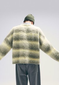 Puszysty, oversize sweter w zielonym kolorze w żółte paski, o miękkiej fakturze. W połączeniu z szarymi spodniami, model ma na sobie zieloną dzianinową czapkę.