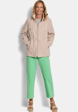 Lächelnde Frau mit blonden Haaren trägt eine beige Jacke mit Reißverschluss und Kapuze, mintgrüne Hose und beige Sandalen mit hohen Absätzen, steht vor einem einfarbigen Hintergrund.