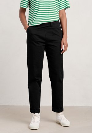 Personne portant un pantalon noir à jambes droites, une chemise rayée verte et blanche, des baskets blanches, debout avec une main dans la poche.