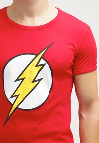 Rotes T-Shirt mit einem großen weißen Kreis und einem gelben Blitzdesign. Das Material erscheint glatt, mit einer taillierten Form und kurzen Ärmeln.