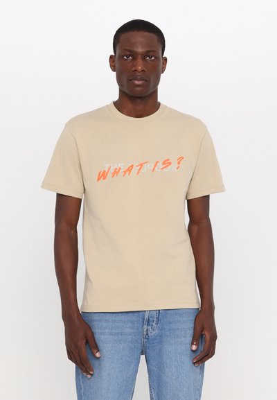 Tricou de bumbac bej, cu un text portocaliu stilizat și îndrăzneț "CE ESTE?" și un model de siluetă albă, cu guler rotund și mâneci scurte.
