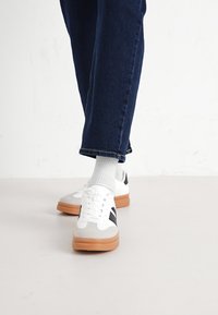 Tenisi albi cu accente gri, dungi negre și o talpă de cauciuc bej. Stilizați cu pantaloni din denim închis și șosete gri.