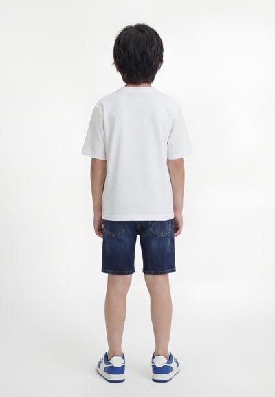 Παιδί που στέκεται με την πλάτη στραμμένη, φορώντας απλό λευκό μπλουζάκι, σκούρο τζιν σορτς και μπλε και λευκά αθλητικά παπούτσια σε λευκό φόντο.