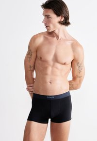 Mustat boxeri alusvaatteet harmaalla vyötärönauhalla, jossa on "SLOGGI"-logo. Sujuva pinta, joustava materiaali, muotoiltu malli ja reiden puoliväliin ulottuva pituus.