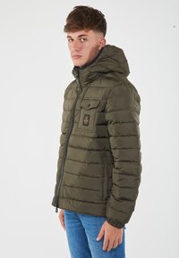 Giacca puffer verde oliva con cappuccio, dotata di chiusura frontale con zip, tasca a toppa sul petto e texture trapuntata.