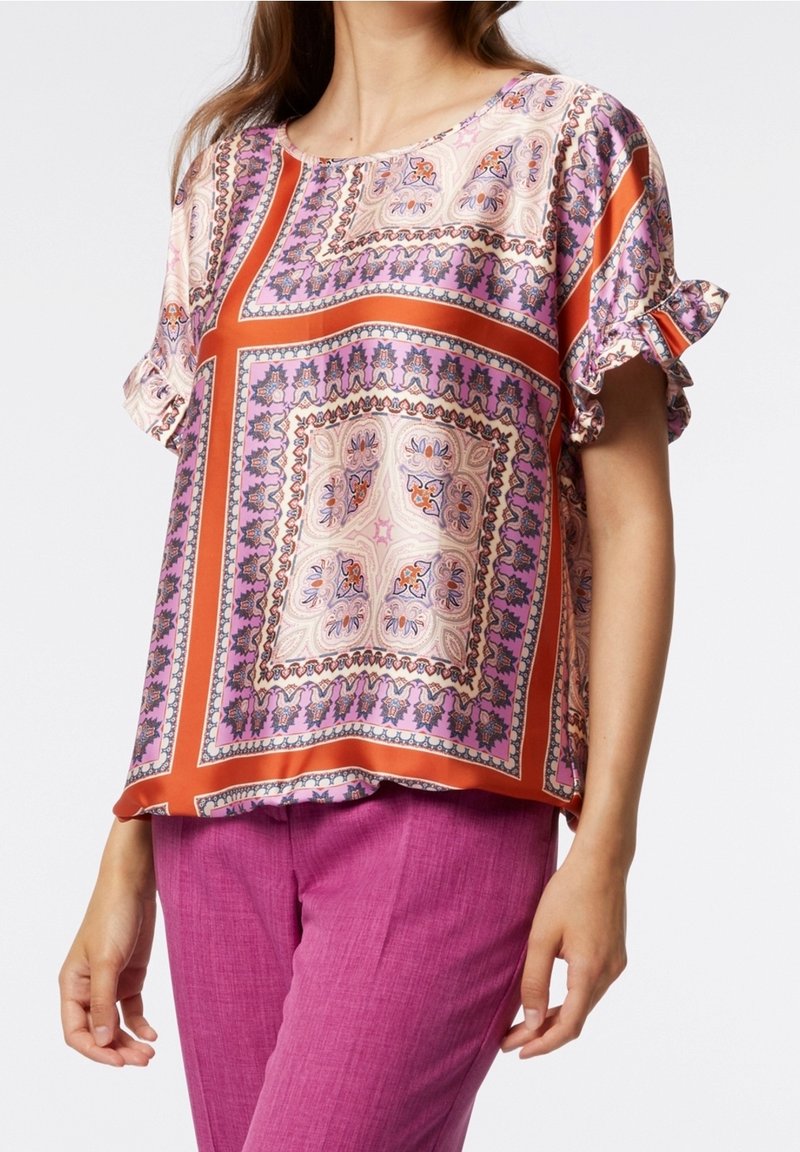 Femme portant un chemisier à motifs avec des dessins géométriques orange et violet et des manches courtes à volants, associé à un pantalon magenta.