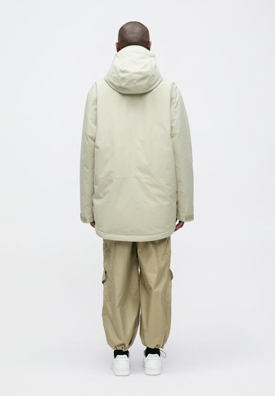 Beige vízálló parka kapucnival, hosszú fazonban, utility-stílusú bézs nadrággal. Fehér sportcipők egészítik ki a szettet.