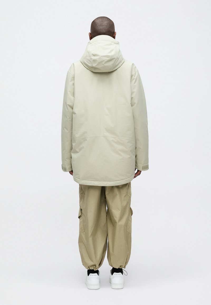 Beiger wasserdichter Parka mit Kapuze, lang geschnitten, kombiniert mit utilitaristischen beigen Hosen. Weiße Sneakers runden das Outfit ab.