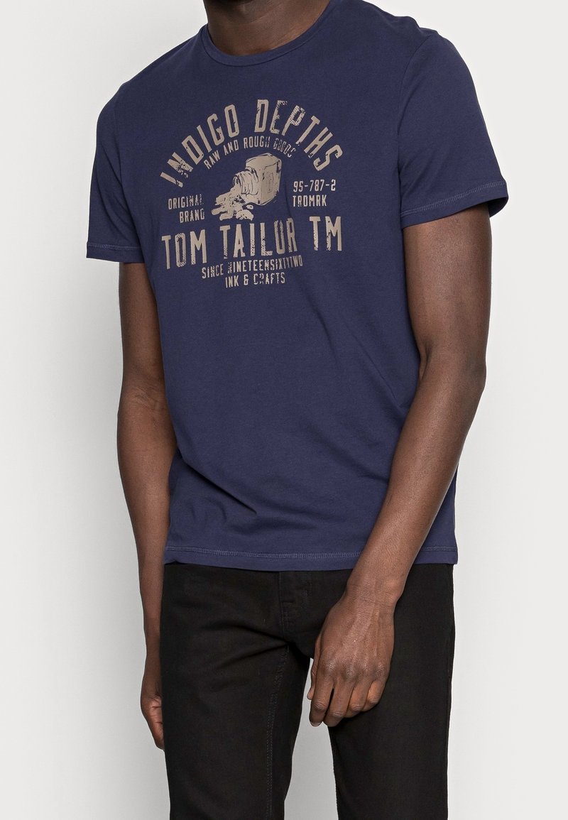 Camiseta de algodón azul marino de manga corta, con una impresión gráfica beige en el frontal que incluye logotipos y texto. Corte casual.