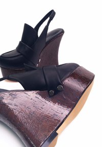 Sandali con zeppa in pelle nera, dotati di un tacco in legno spesso e strutturato, una larga fascia che attraversa il piede e piccoli dettagli metallici.