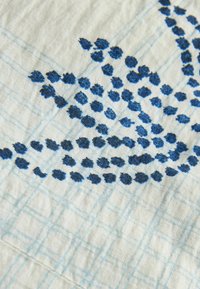 Tissu blanc avec des lignes de quadrillage bleu clair et un motif brodé de points bleu foncé regroupés formant une forme de branche.
